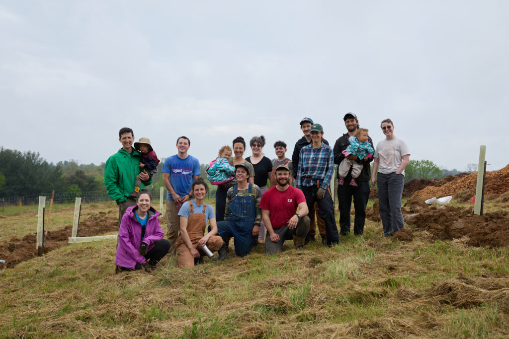 A group of people in two rows, the back row standing and the front row kneeling, in work clothes on a grassy stretch of land. Three of the adults are holding small children. Everyone is smiling, and the sky is grey.