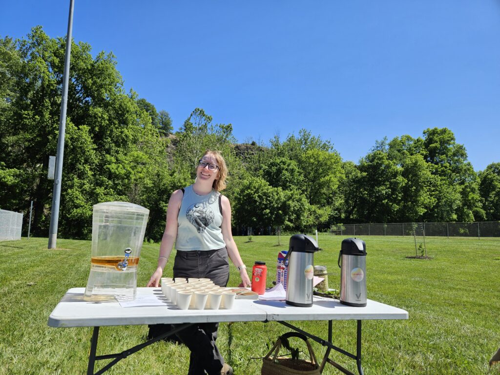 A woman standing behind a folding table with a large beverage dispenser with amber liquid in it, and rows of small paper cups next to it. There are two hot beverage carafe dispensers next to the paper cups. She is standing in front of a line of trees under a clear blue sky, in a mowed park field. 