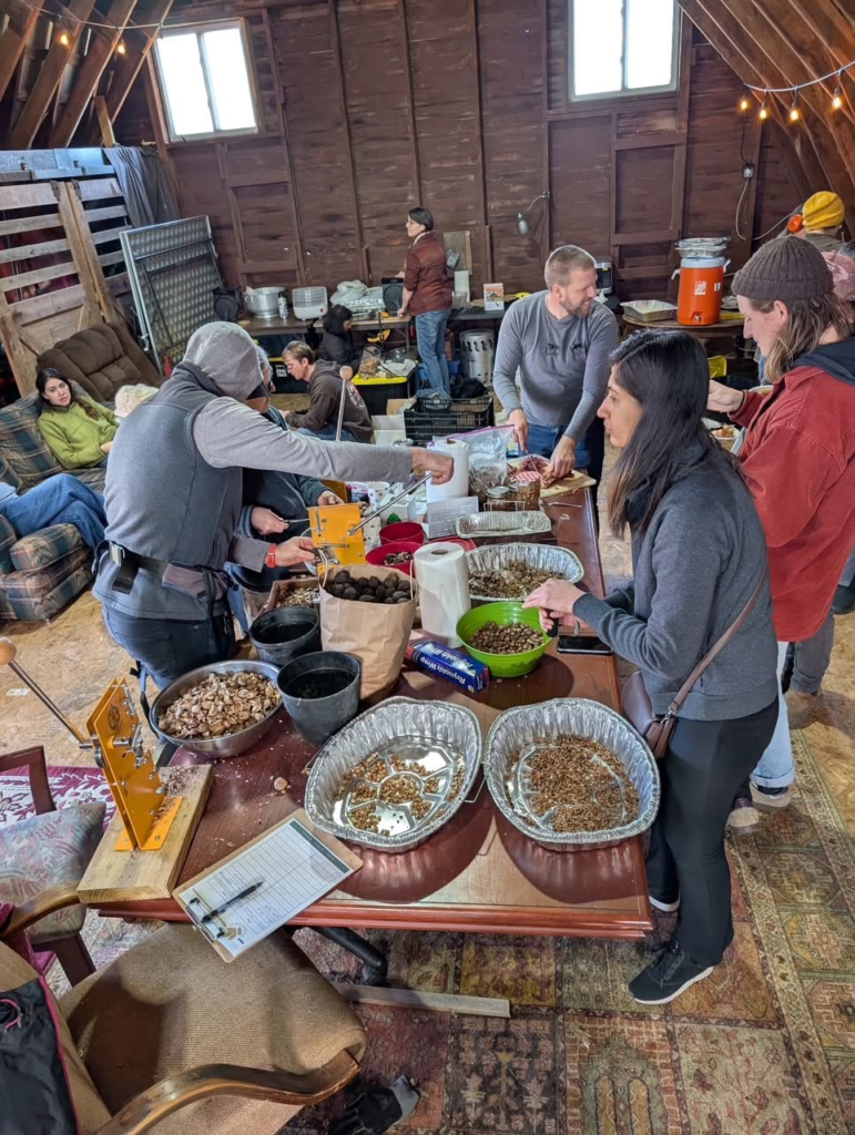 An indoor gathering inside of a barn with a long table covered in buckets and tins filled with tree foods, surrounded on all sides by people who are serving themselves from the different containers.
