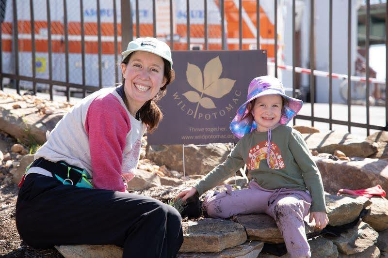 A mother and her child sitting on a wall of stones smiling at the camera, on either side of a brown sign that reads: Wild Potomac with a large leaf logo.