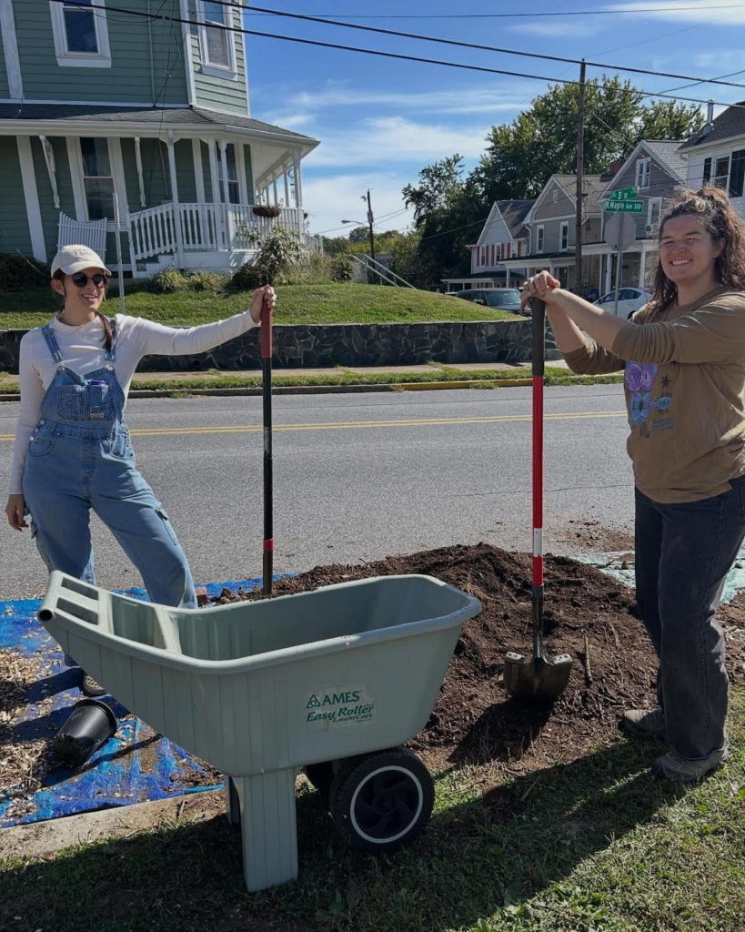 Two people standing next to a road, each with a large shovel in their hands, posing next to a hole that they have dug with a wheelbarrow in front of them.