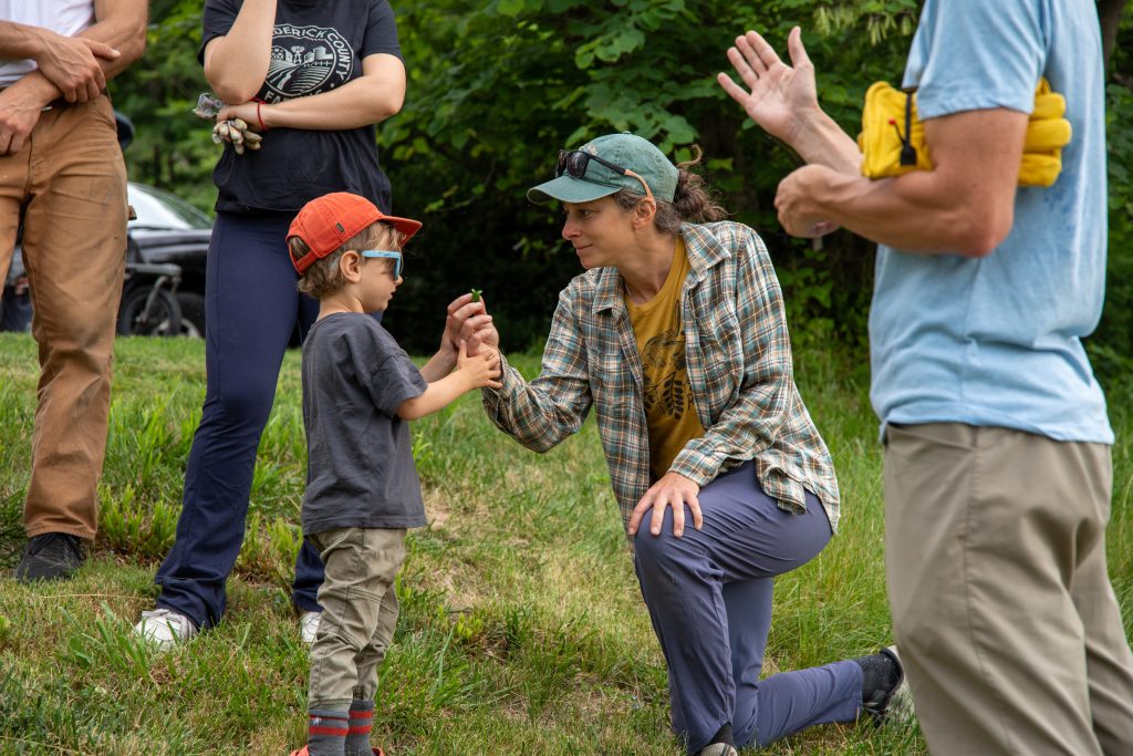 A mother kneeling down on the grass holding up a plant and her small child standing in front of her, holding her hand with both of his as they both look at the plant.