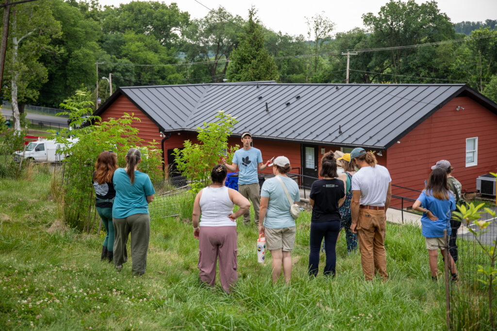 A small crowd of people gathered in a circle in front of a man who is gesturing with his hands, talking to them. They are standing outside on the grass in front of a red building.